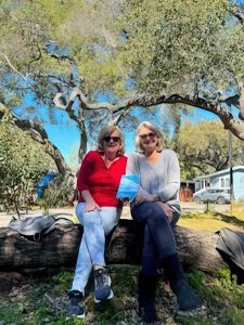 Suzanne and Patty sitting outside on a log holding their book